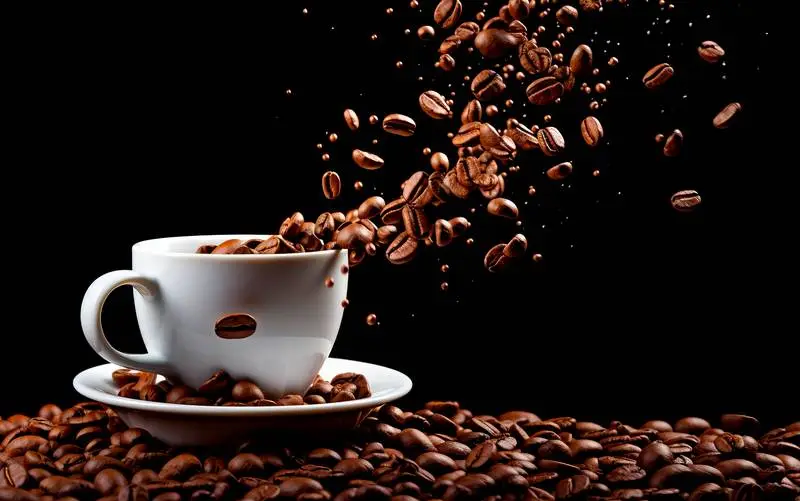 White Cup on a Surface of Coffee Beans and Other Beans Falling on a Black Background
